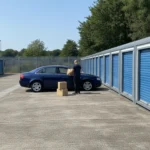 Modular storage units in a concrete car park with a person unloading boxes from a car.